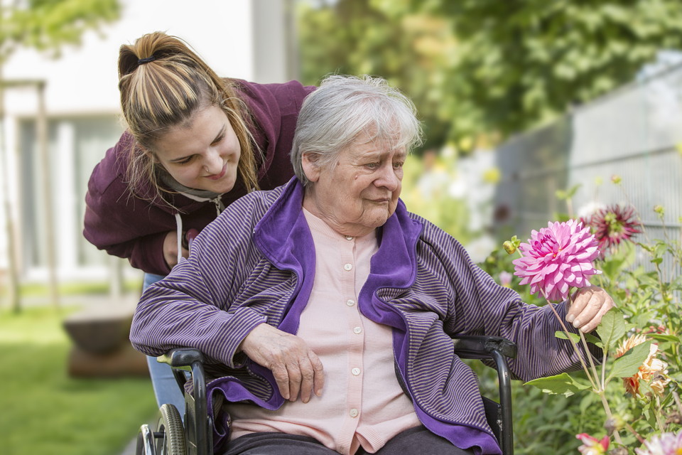 Pflegerin mit einer Heimbewohnerin im Rollstuhl beim Betrachten einer Blume
